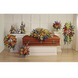 Bright multicolor funeral floral set with casket spray, wreath, and standing sprays around a wooden casket.