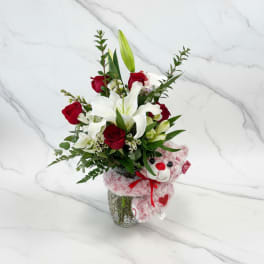 Red roses and white lilies in a glass vase with a pink teddy bear