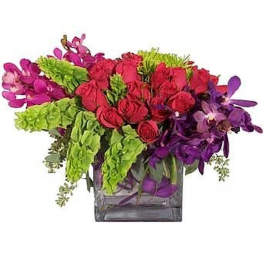 Low arrangement of red roses, green bells of Ireland, and purple orchids in a glass cube vase
