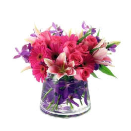 Low arrangement of pink gerbera daisies, roses, and lilies with purple orchids in a clear glass vase