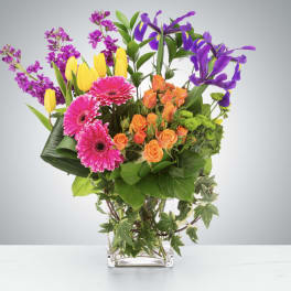Mixed bouquet with pink gerbera daisies, orange roses, yellow tulips, and purple irises in a glass vase