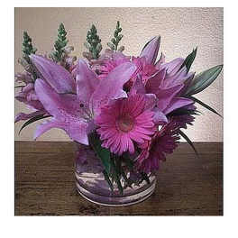 Low arrangement of pink lilies and gerbera daisies in a clear glass vase on a wooden surface