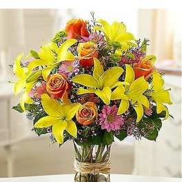 Yellow lilies, orange roses, and pink daisies arranged in a clear glass vase with pebbles