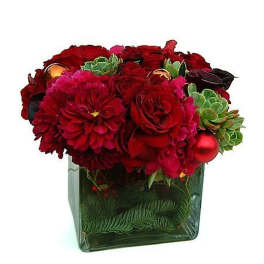 Low red and green floral arrangement with roses, dahlias, succulents, and ornaments in a clear glass cube vase