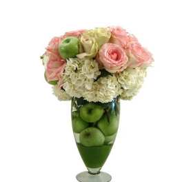 Tall glass vase filled with green apples topped with white hydrangeas and pink roses