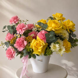 Pink carnations and yellow roses arranged in a white bucket vase with a pink ribbon.