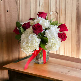 Arrangement of red roses and white hydrangeas in a clear glass vase with a red ribbon.