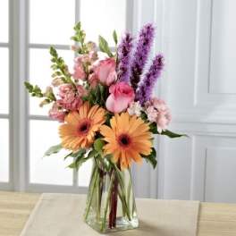 Mixed bouquet of orange gerbera daisies, pink roses, and purple spikes in a clear glass vase