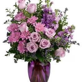 Lavender roses and pink mums arranged with purple blooms in a purple glass vase