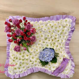 Pillow-shaped white mum tribute with purple ribbon edging, roses, and a blue hydrangea cluster.