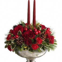 Low red rose and carnation centerpiece with holly, pinecones, and two tall red taper candles in a silver bowl.