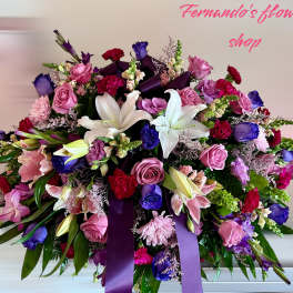 Large mixed arrangement of white lilies, pink roses, purple blooms, and red carnations with a dark purple ribbon