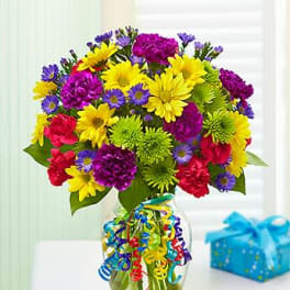 Bright mixed bouquet of yellow daisies, carnations, and green mums in a glass vase with curling ribbons.