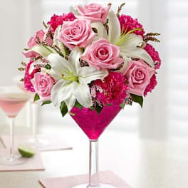 Pink roses, white lilies, and bright pink blooms arranged in a martini glass vase with pink filler.