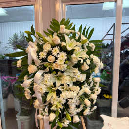 Large white funeral spray on an easel with lilies, roses, and ribbon