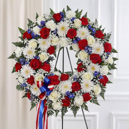 Standing wreath of red roses, white mums, and blue flowers with a patriotic ribbon on an easel.