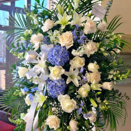 Tall standing spray of white roses, lilies, and blue hydrangeas with palm fronds on an easel.