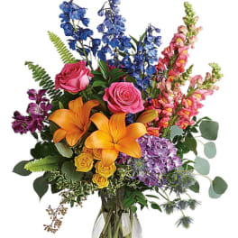 Colorful bouquet with roses, lilies, and tall blue and pink blooms in a glass vase