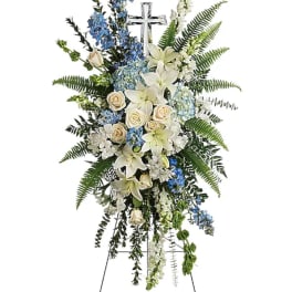 Standing floral spray with white lilies, cream roses, and blue flowers on an easel cross