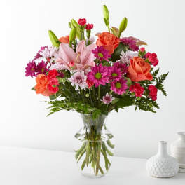 Mixed bouquet of pink lilies, orange roses, and purple daisies in a glass vase