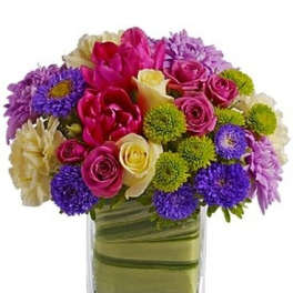 Mixed bouquet of pink, purple, and cream flowers in a clear square vase