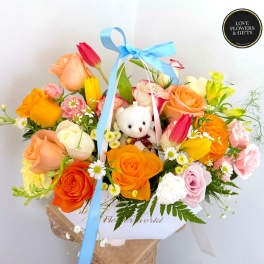 Bright mixed arrangement of roses and tulips with a small white teddy bear in a white box.