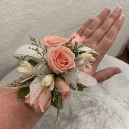 Wrist corsage with peach roses, white buds, and sparkly white ribbon on an arm.
