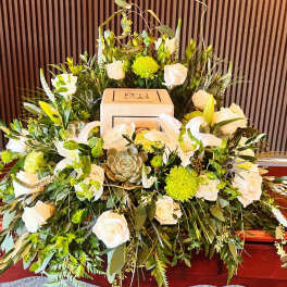 Cremation urn surrounded by white roses, lilies, green mums, and a succulent on a wooden casket
