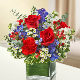 Bouquet of red roses, red carnation, blue flowers, and white daisies in a green-lined glass cube vase