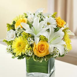 Yellow and white lilies, roses, and daisies arranged in a square glass vase