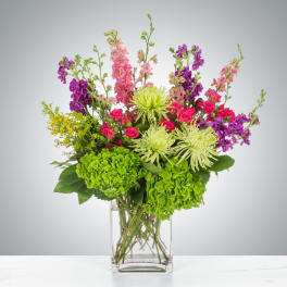 Tall mixed arrangement with green hydrangeas, lime mums, pink roses, and purple stock in a clear glass vase