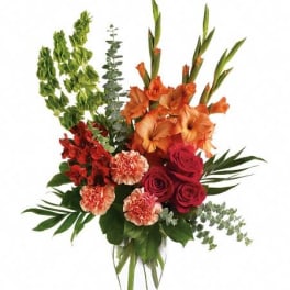 Tall arrangement with orange blooms, red roses, peach carnations, and green bells in a clear glass vase