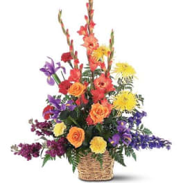 Tall mixed flower arrangement with orange gladiolus, roses, yellow mums, and purple blooms in a wicker basket