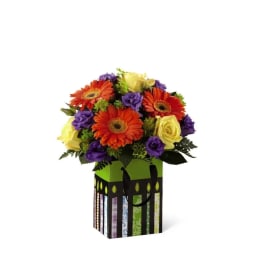 Bouquet of orange gerbera daisies, yellow roses, and purple flowers in a gift bag