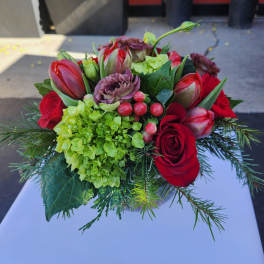 Short arrangement of red roses, tulips, green hydrangea, berries and lisianthus in a glass vase with evergreen sprigs.