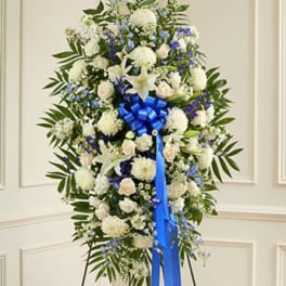 Standing spray of white lilies, roses, and blue flowers with a blue bow on an easel.