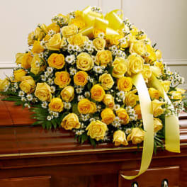 Yellow rose and white daisy casket spray with a large yellow ribbon bow on a wooden casket