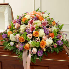 Colorful casket spray with orange roses, pink lilies, white mums, and purple flowers on a wooden casket