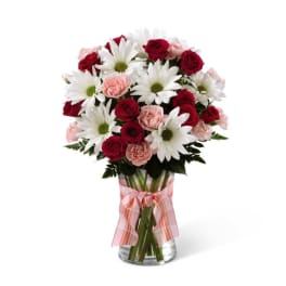 Tall bouquet of red roses, white daisies, and pink carnations in a clear glass vase with a pink ribbon.