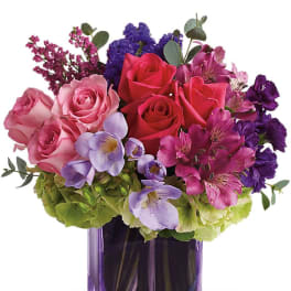 Compact arrangement of pink and red roses with purple blooms in a purple glass cube vase