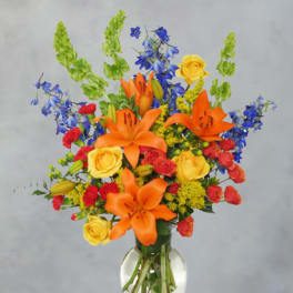 Bright mixed bouquet with orange lilies, yellow roses, and blue flowers in a glass vase