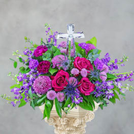 Purple and pink floral arrangement with a clear cross topper in a pedestal vase