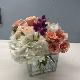 Pink roses and white hydrangeas in a clear square vase with purple orchids