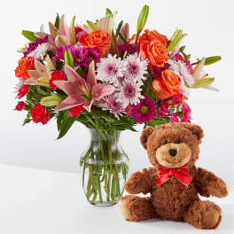 Mixed bouquet in a glass vase beside a teddy bear with a red bow
