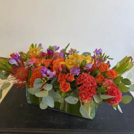 Low rectangular arrangement of orange, yellow, and purple flowers in a glass vase