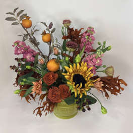 Autumn floral arrangement with sunflowers, roses, pink blooms, and oranges in a green vase