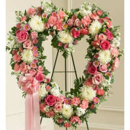 Heart-shaped standing wreath of pink roses, carnations, and white flowers with a pink ribbon bow.