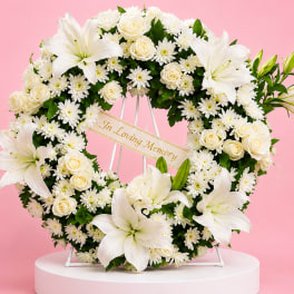 Heart-shaped white floral wreath with lilies, roses, and daisies on a stand
