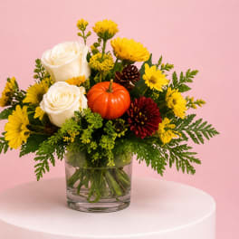 White roses and yellow flowers arranged in a glass vase with a small pumpkin