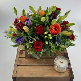 Mixed bouquet with roses, lilies, and purple blooms in a wooden box beside a candle